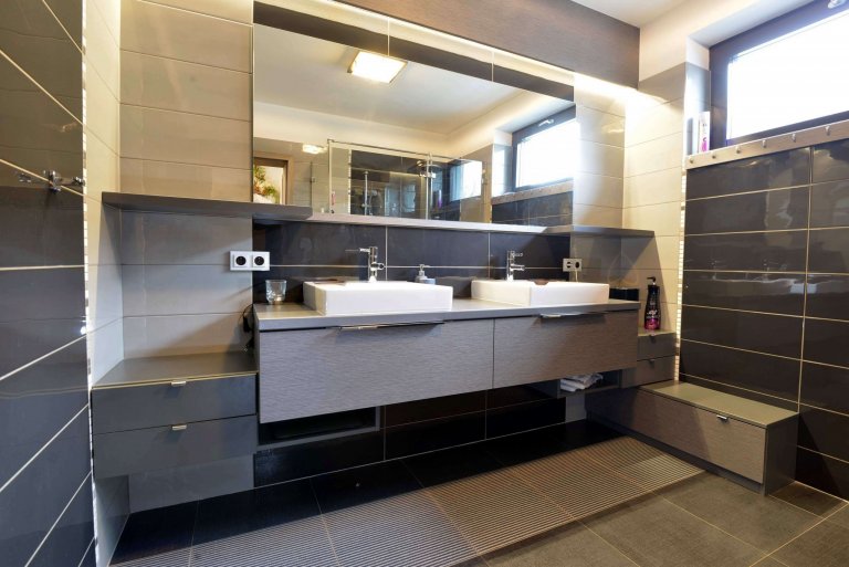 Koupelna pro rodiče. Vše je v odstínech šedé a černé. Toto je pohled na koupelnový nábytek s dvěma umyvadly, každý z páru má svojí stranu. Sestavu doplňuje velkoplošné zrcadlo a lišta s osvětlením LED. 