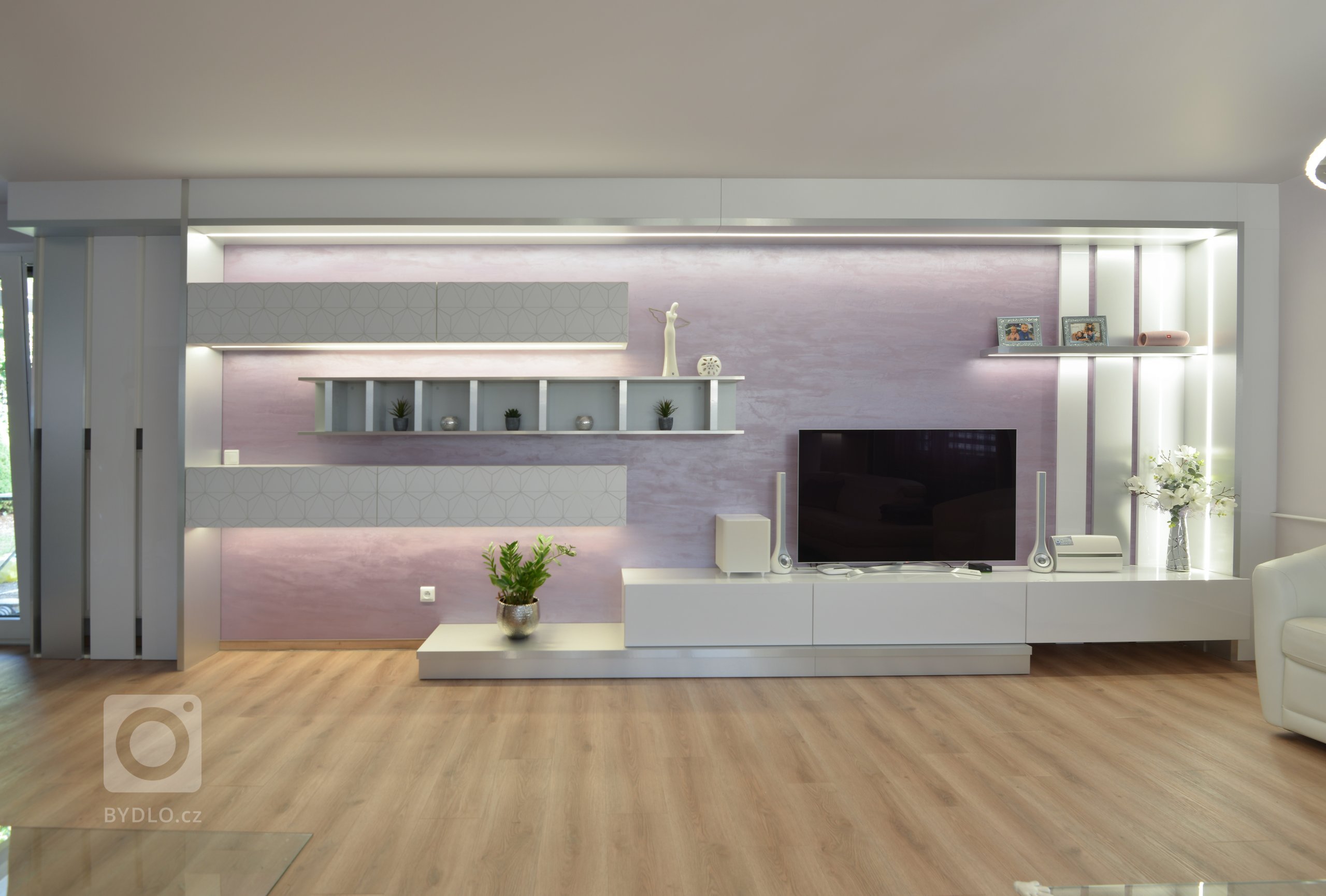 Pohledová stěna pro hlavní sestavu nábytku je uměle orámovaná v kombinaci bílé lesklé a kovolaminátu z kartáčovaného hliníku se zabudovaným osvětlením. Závěsné skřínky jsou z bílé matné s potiskem overface. Regálek je z kovolaminátu. Skřínky jsou výklopné a sklopné, dole jsou hluboké zásuvky. 