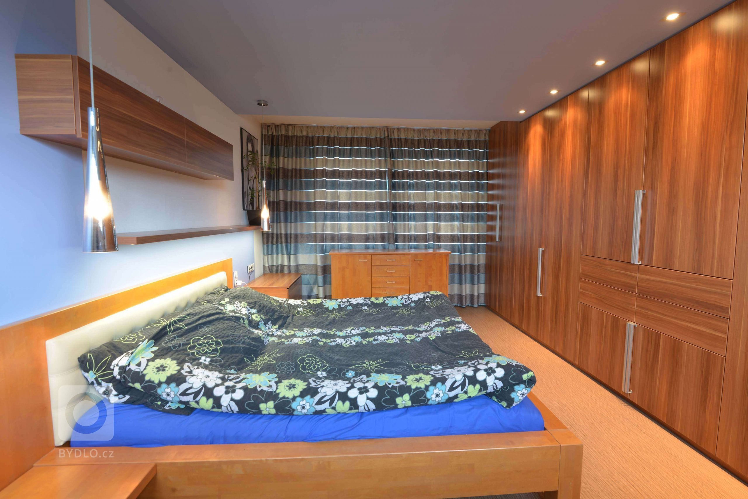 Za postelí je předstěna, která prochází přes strop ke skříním a má skryté osvětlení LED. Na předstěně je otevřená i zavřená knihovna. 