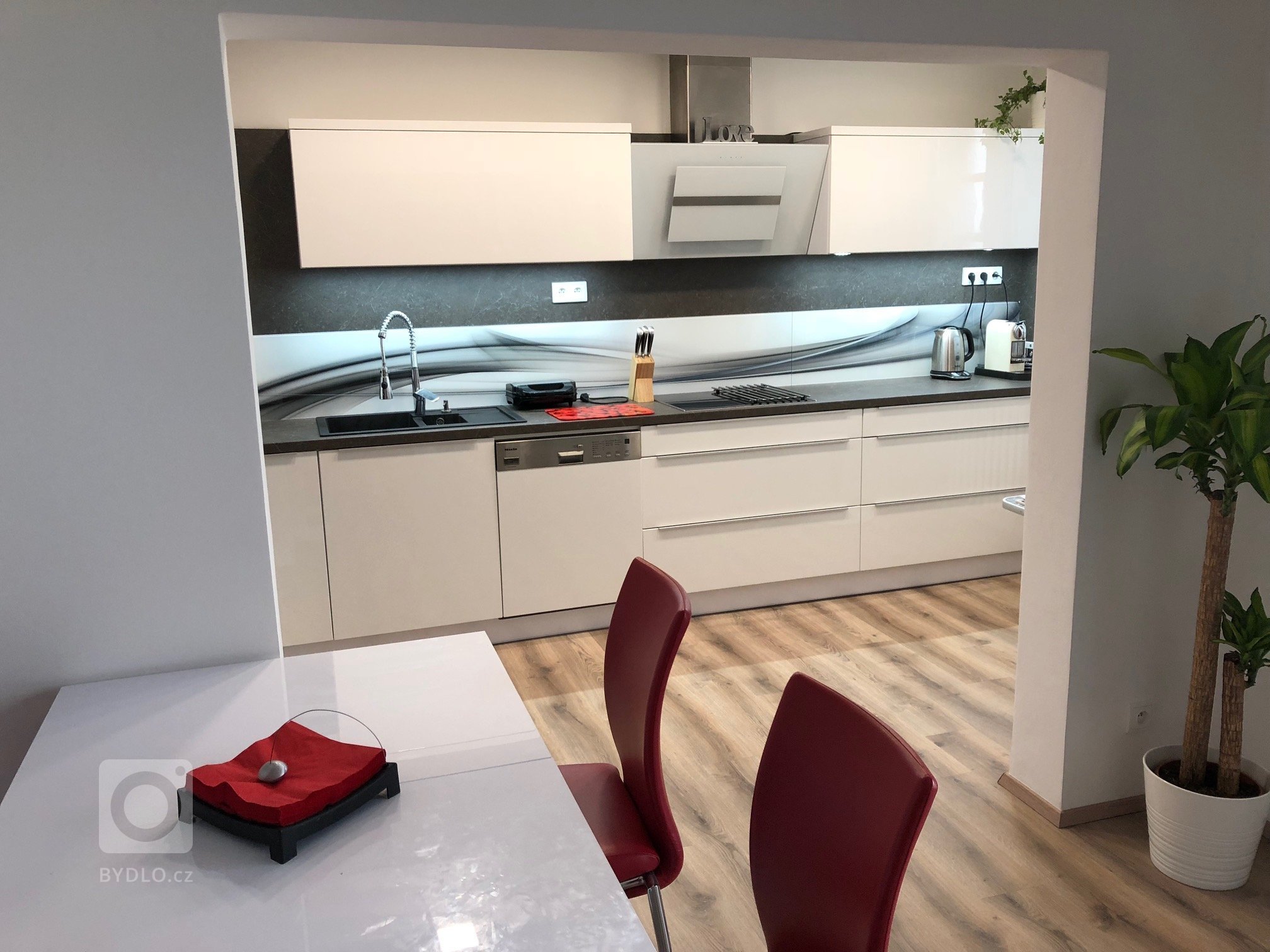 Kuchyňká linka v bílém vysokém lesku s grafosklem, osazená spotřebiči Bosch a doplněná příruční stolovou deskou zabudovanou do skříňkové sestavy protilinky.   