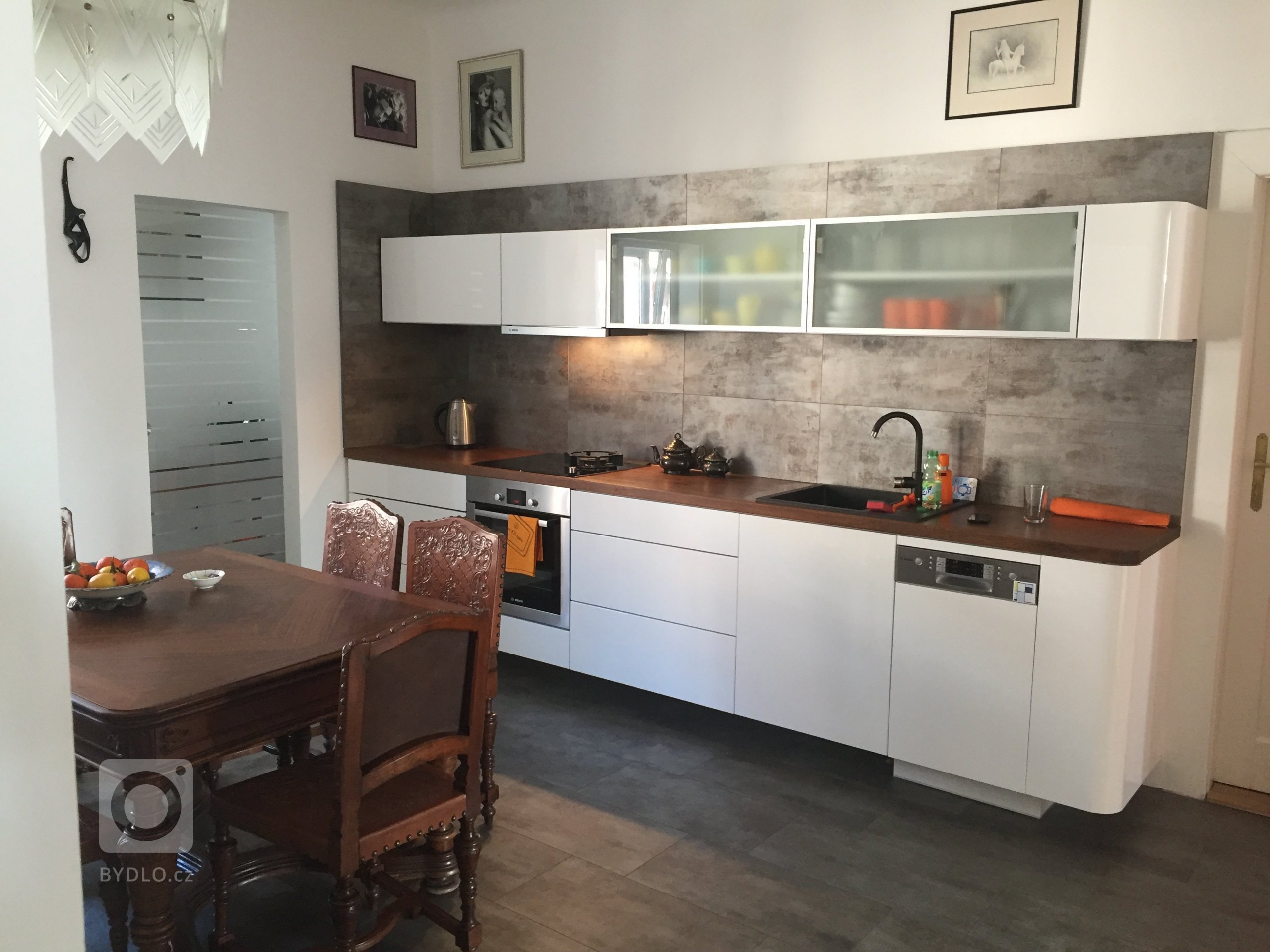 Kuchyňská linka v bílém vysokém lesku doplněná masivní pracovní deskou. V kuchyni je umístěn námi renovovaný starožitný stůl s židlemi a konferenční stolek.
