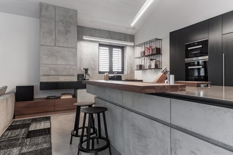 Subtilní pracovní deska z umělého kamene se dostává do kontrastu s masivní barovou deskou z ořechu, s místy k sezení pro dvě osoby.