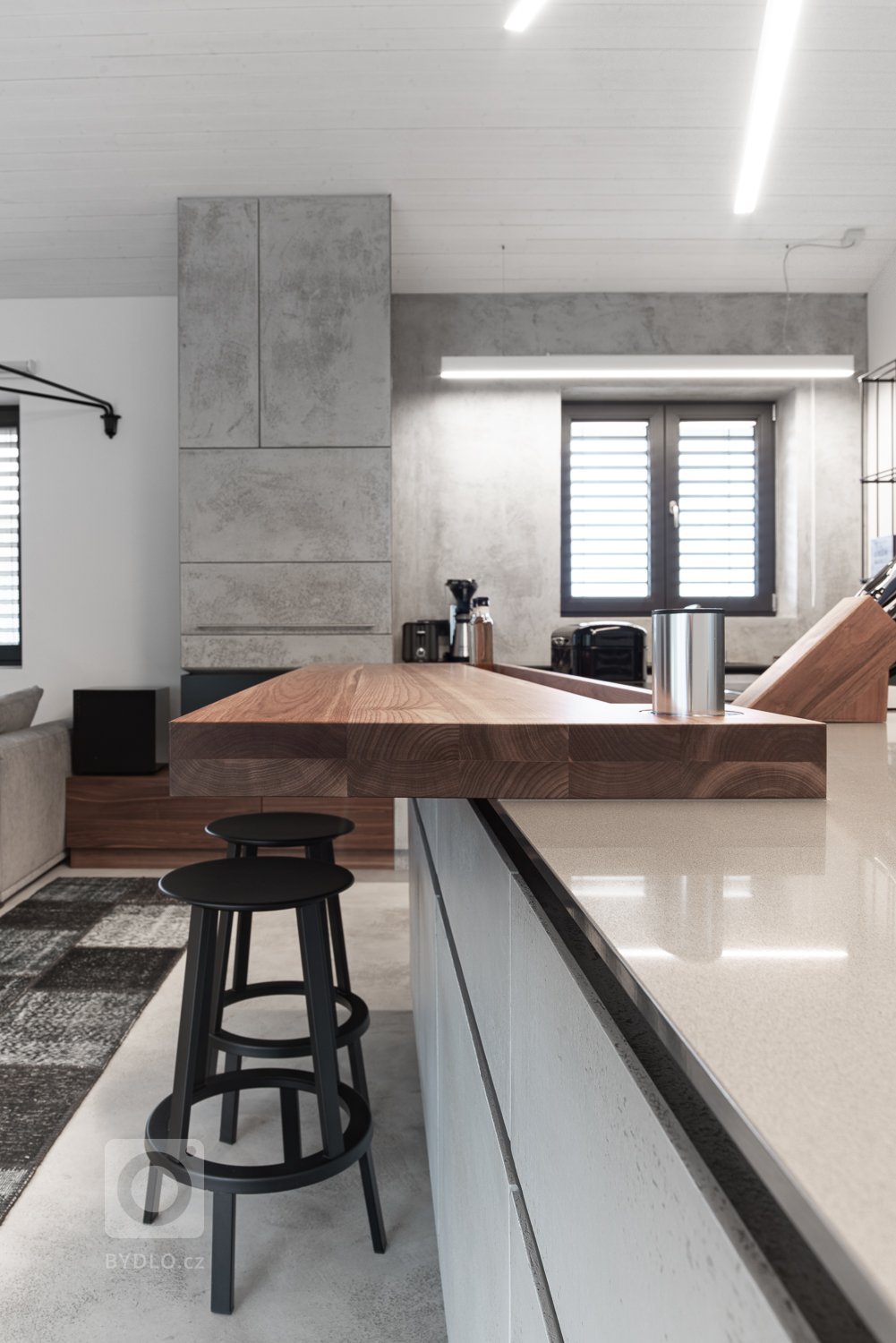 Subtilní pracovní deska z umělého kamene se dostává do kontrastu s masivní barovou deskou z ořechu, s místy k sezení pro dvě osoby.