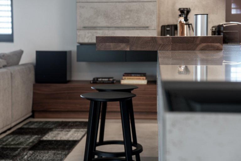 Subtilní pracovní deska z umělého kamene se dostává do kontrastu s masivní barovou deskou z ořechu, s místy k sezení pro dvě osoby. 